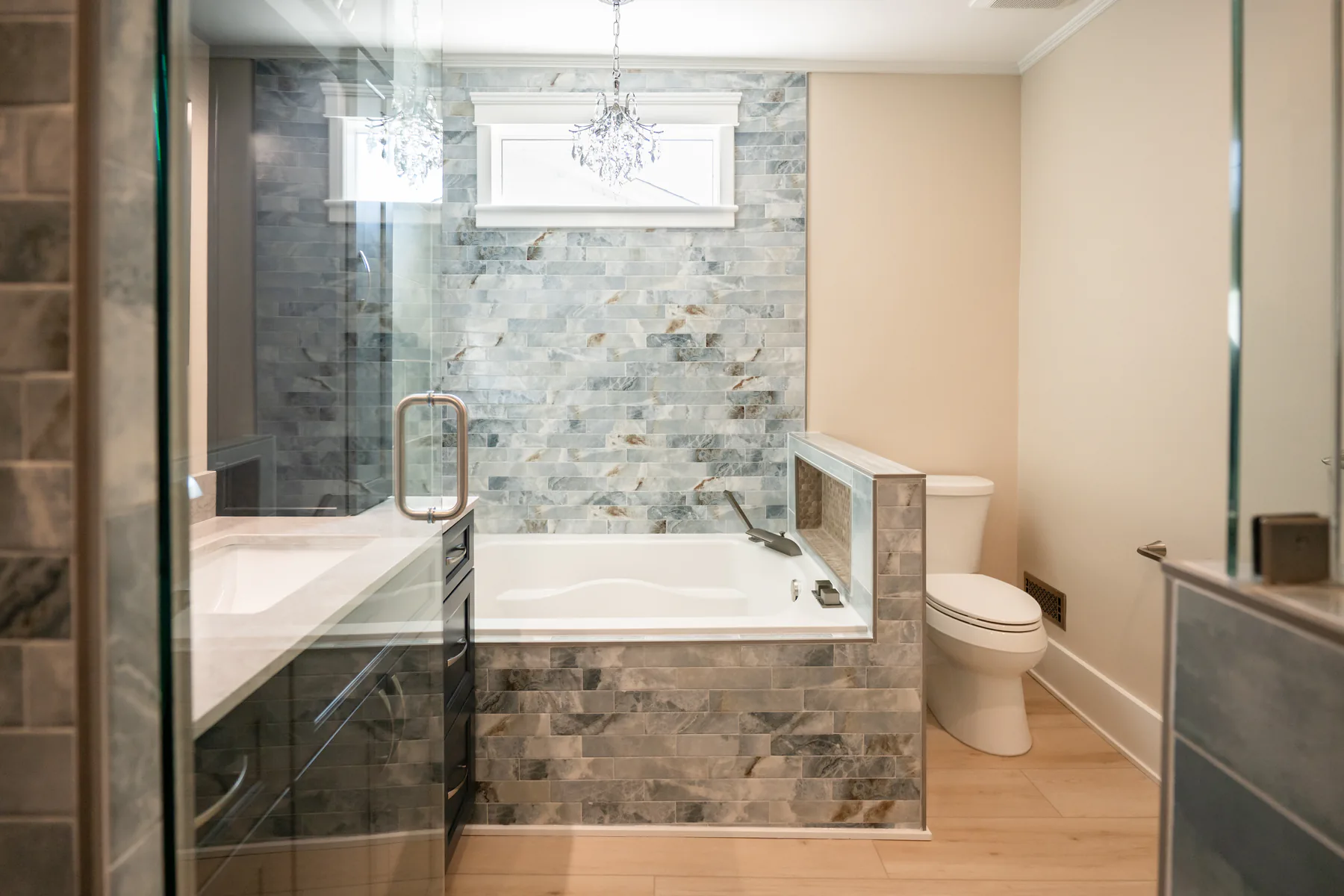 Completed bathroom remodel with blue-gray tile, bathtub, toilet, glass shower, and light wood-look flooring
