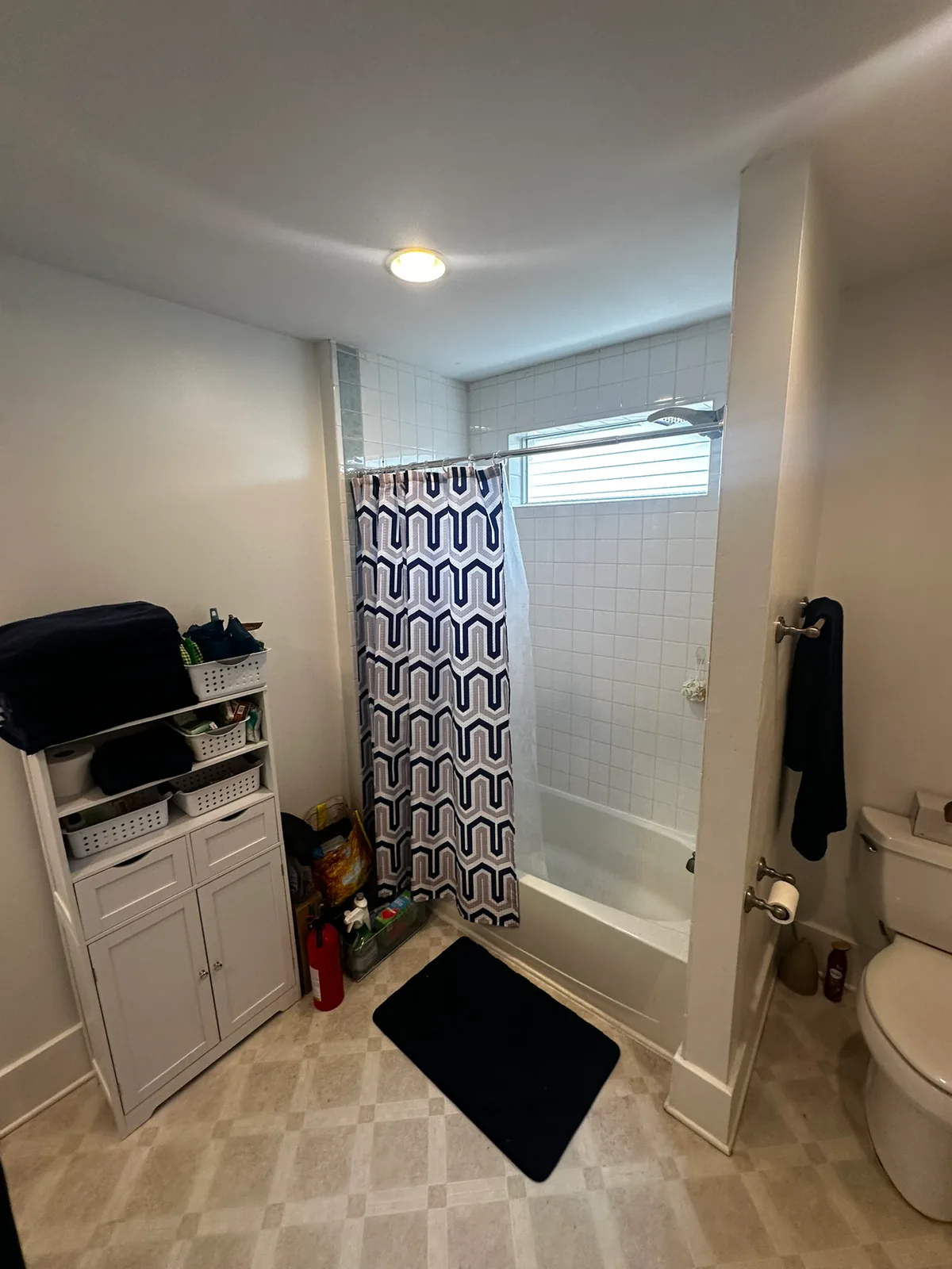 Bathroom before remodeling with white tiled tub surround, shower curtain, toilet, freestanding storage, and light vinyl flooring