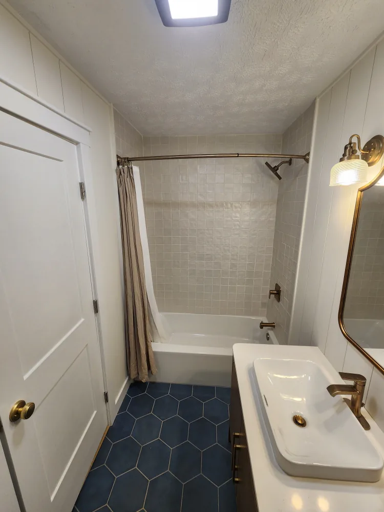 Completed bathroom remodel with white bathtub, light tiled surround, white countertop sink, brass fixtures, and navy hexagonal floor tile